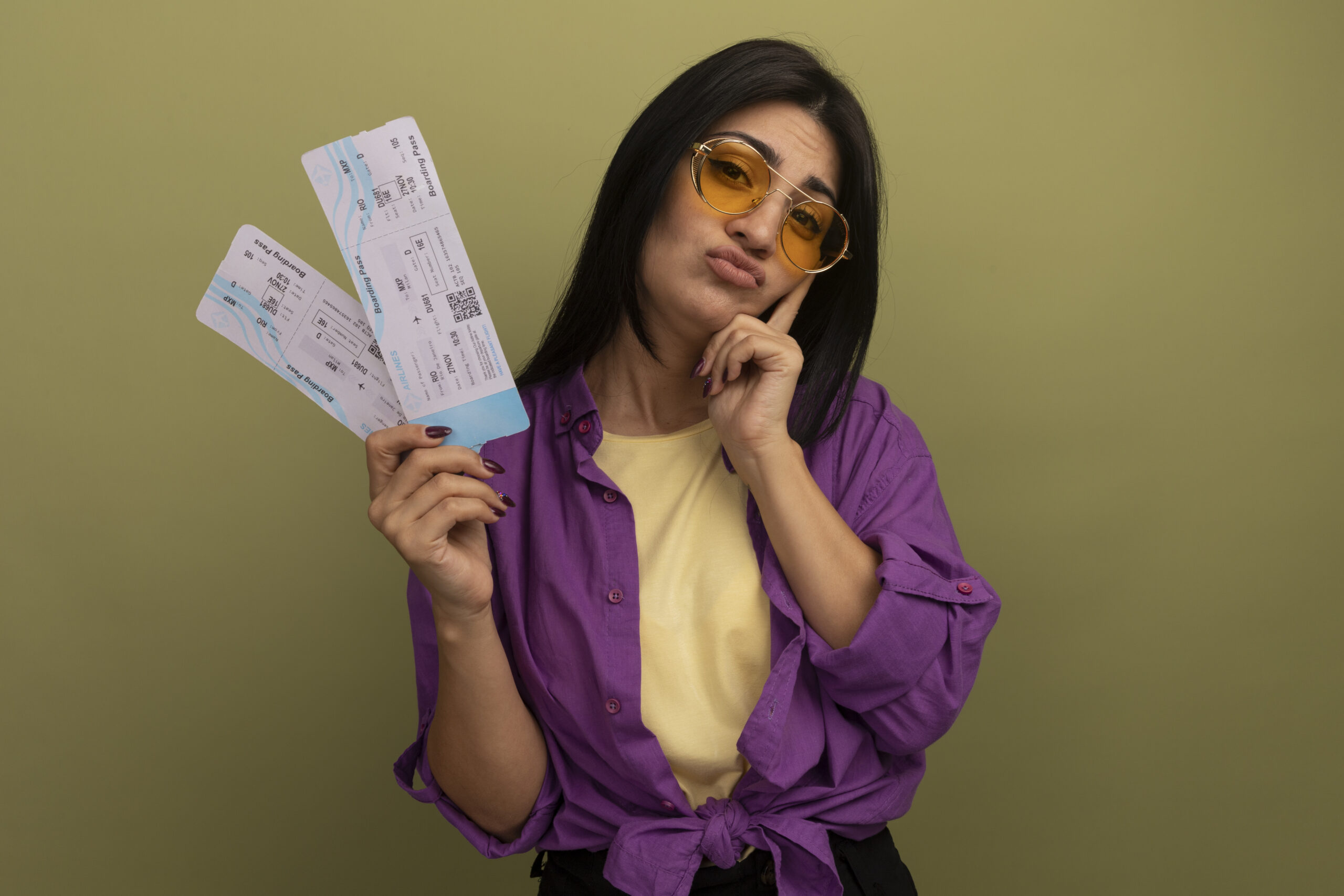 unpleased pretty brunette caucasian girl in sun glasses puts finger on face and holds air tickets isolated on olive green background with copy space
